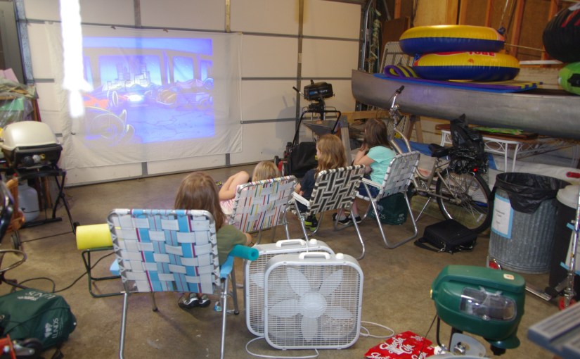 Kids in lawn chairs watching a movie projected on the door of a garage.